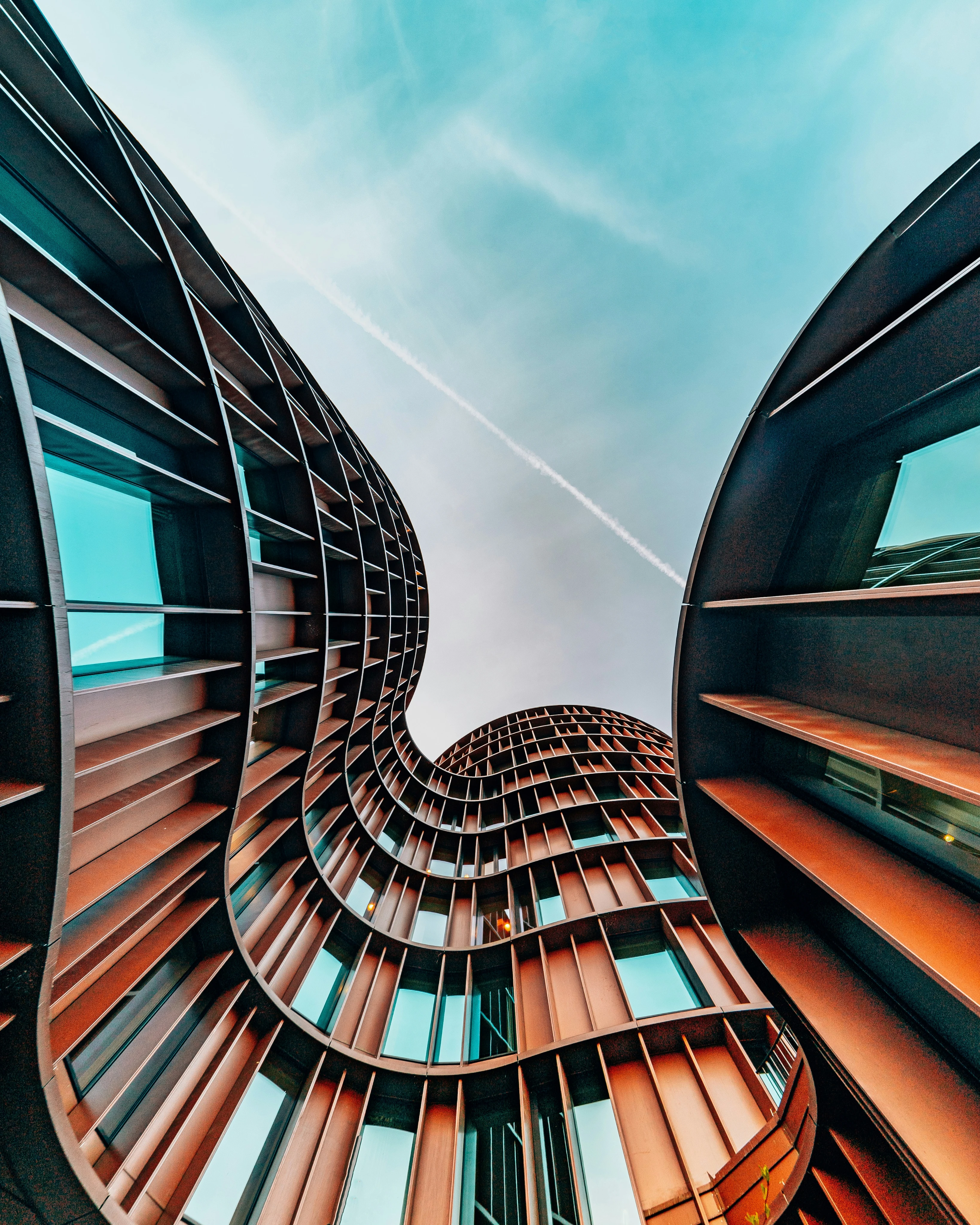 Curved modern brown building with blue windows against a partly cloudy sky with a contrail.
