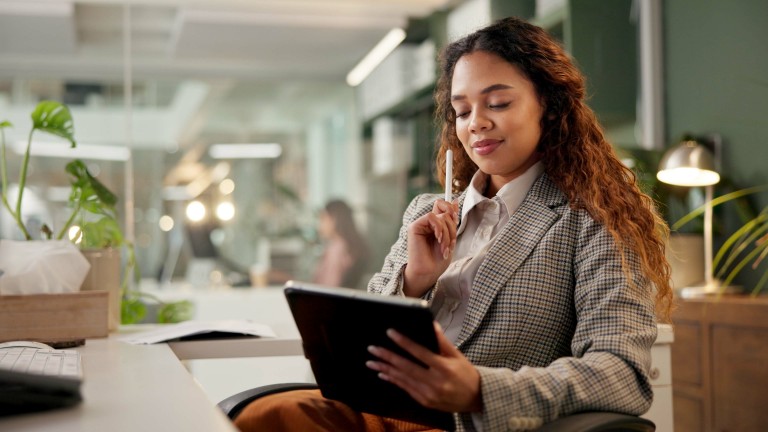 Young woman in office thoughtfully holding a pencil and looking at a tablet.