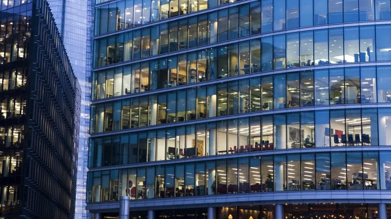 Curved office building with many glass windows showing illuminated workspaces inside at dusk.