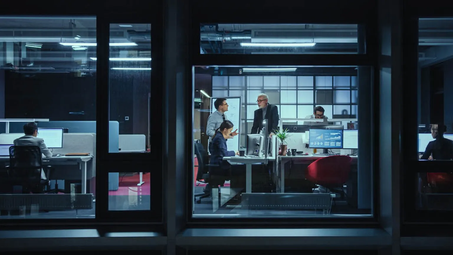Office workers at night, some seated at desks on computers, others standing and discussing inside a glass-walled room.
