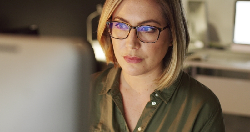 Woman with glasses focused on a computer screen in a softly lit office environment.