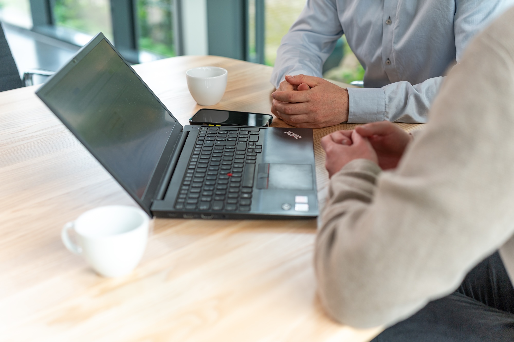 Zwei Personen sitzen an einem Holztisch mit einem Laptop, einem Smartphone und zwei weißen Tassen.