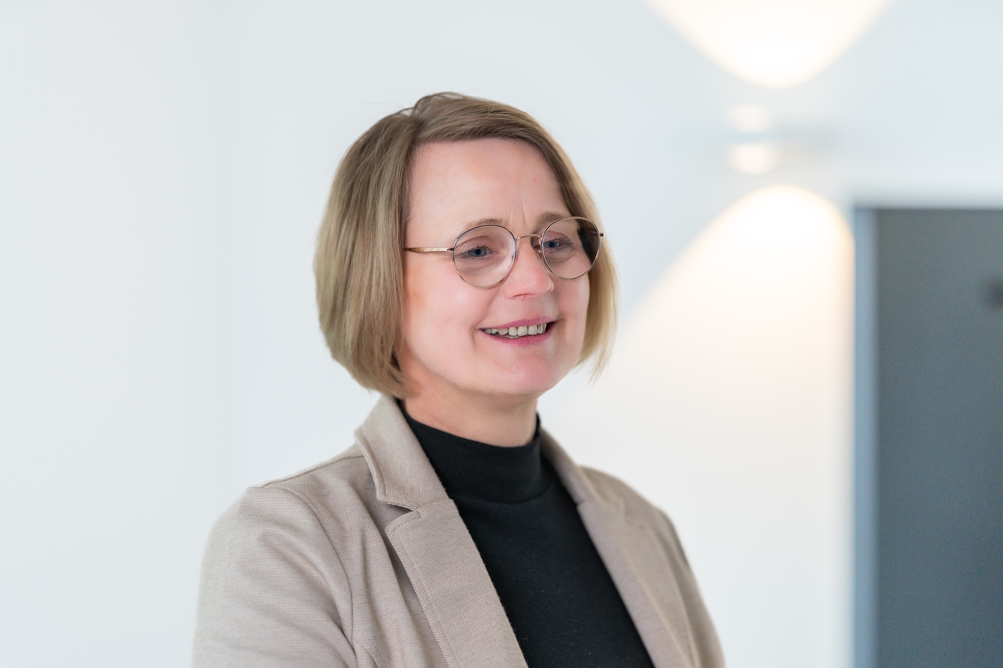 Lächelnde Frau mit kurzen blonden Haaren, Brille, schwarzem Rollkragenpullover und beiger Jacke vor hellem Hintergrund.