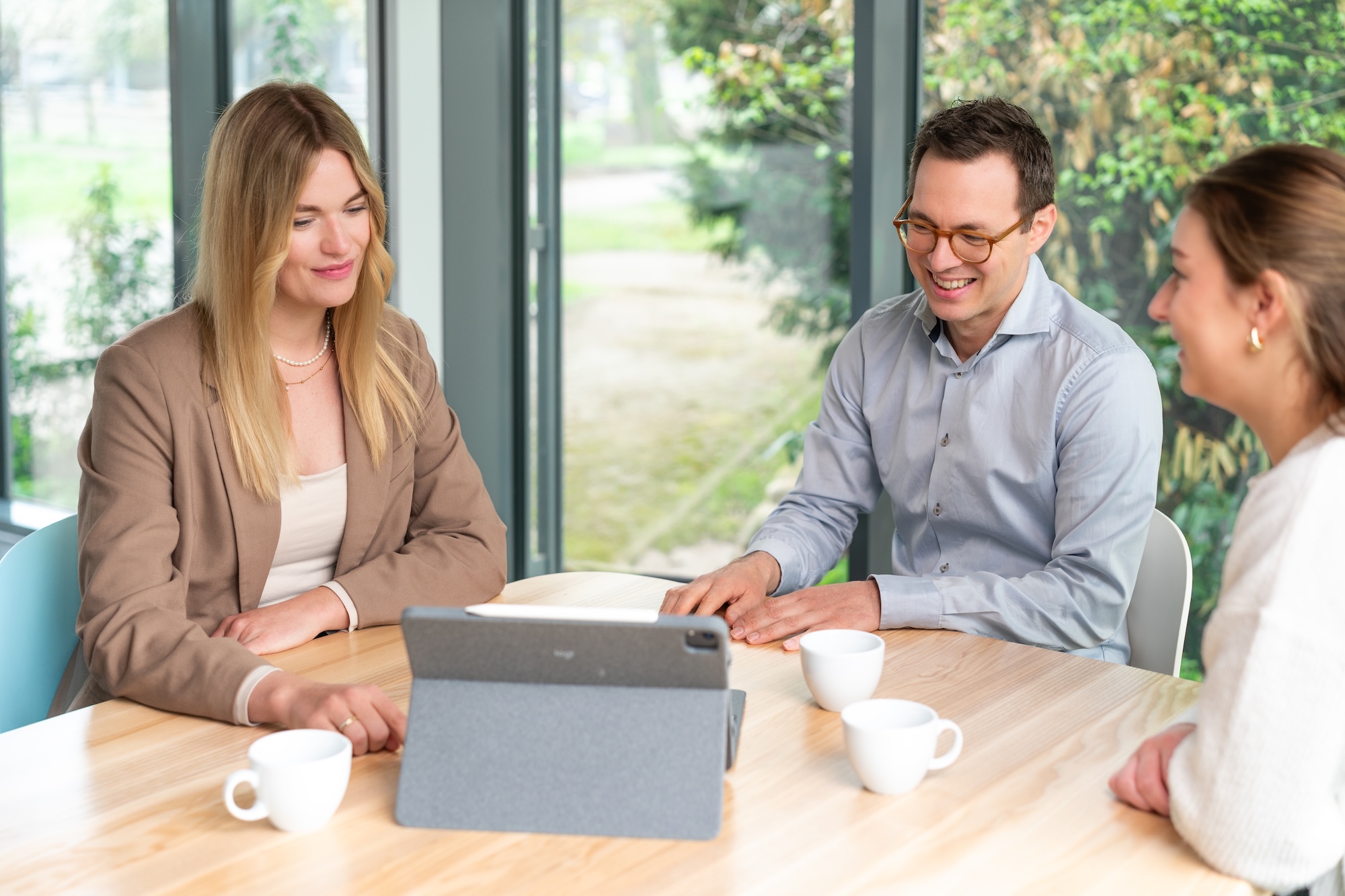 Drei Personen sitzen an einem Holztisch vor einem Tablet und trinken aus weißen Tassen in einem hellen Raum mit großen Fenstern.