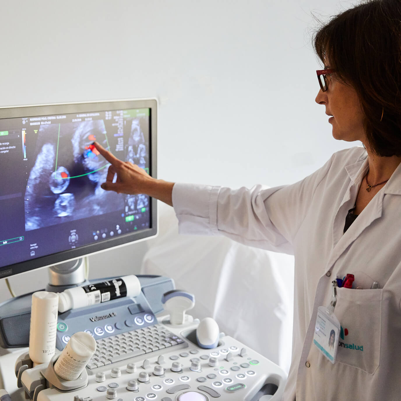 A doctor in a white lab coat points at an ultrasound image on a monitor, situated in a clinical setting with an ultrasound machine prominently in view.