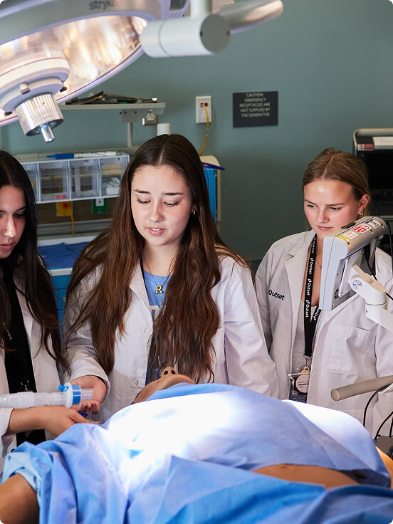Three students in white lab coats observe a medical procedure. One person adjusts a tool on a simulated patient in a clinical setting, under bright surgical lights.