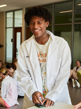 A future medical student wearing a white lab coat smiles while standing near a table. In the background, several people in similar attire sit, conversing in a modern indoor space.
