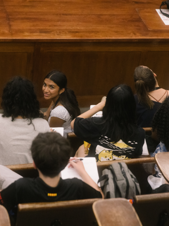 A group of future medical students sits in an auditorium, taking notes. One student smiles and looks back towards the camera. The wooden seating adds a classic lecture hall feel.