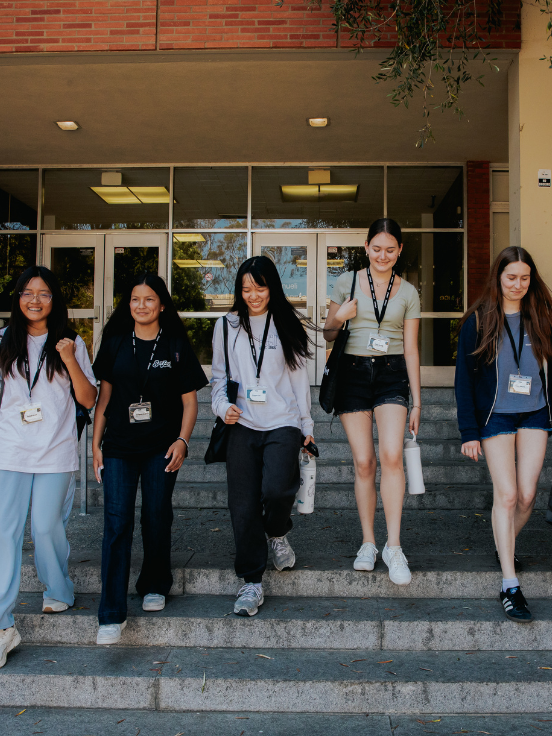 Five students walking down steps, wearing lanyards and casual attire. They appear to be leaving a building with large glass doors and brickwork, under a shaded entrance with overhead lights.