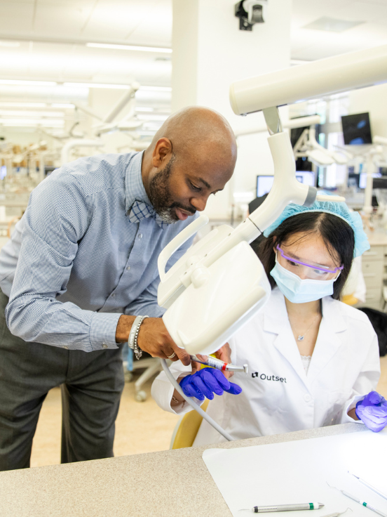 Instructor assists student with dental equipment. Student, wearing protective gear, is seated in a laboratory. Instructor leans over, demonstrating a technique. Text on lab coat reads "Outset."