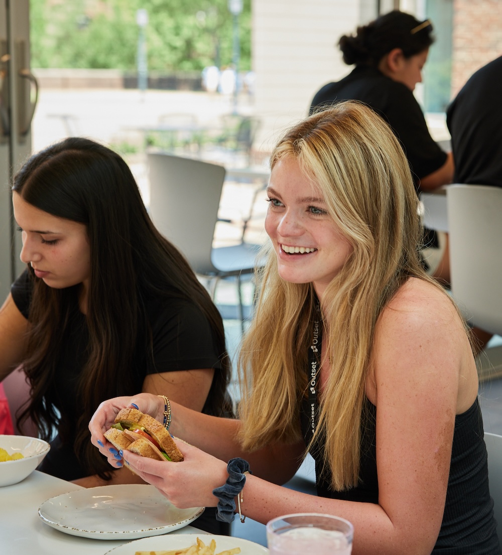 Two students sit at a table in a bright indoor setting, one smiling and holding a sandwich, while the other focuses on her food.