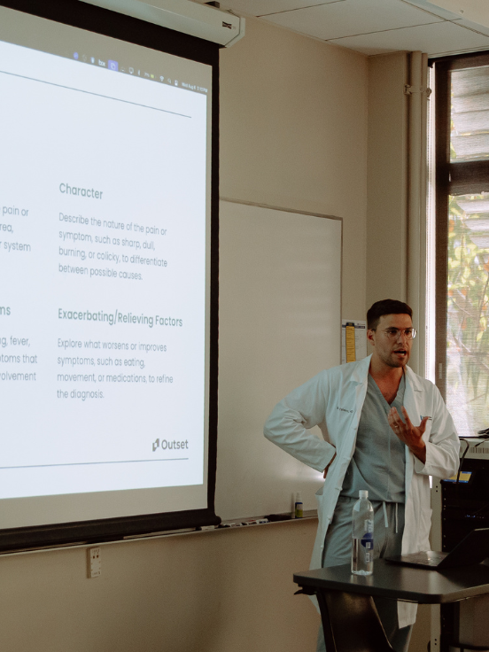 A doctor talks beside a projected presentation slide, detailing medical symptoms and factors, in a classroom with a window. Items on the table include a water bottle and sanitizer. Text on slide:
- "Character
Describe the nature of the pain or symptom, such as sharp, dull,
burning, or colicky, to differentiate between possible causes.
- Exacerbating/Relieving Factors
Explore what worsens or improves symptoms, such as eating, movement, or medications, to refine the diagnosis.
- Outset"
