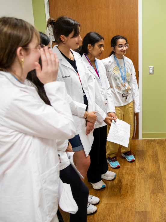 Five future medical students wearing lab coats stand in a hallway, holding papers, appearing to listen attentively. The setting includes a wooden floor, green walls, and a closed door.