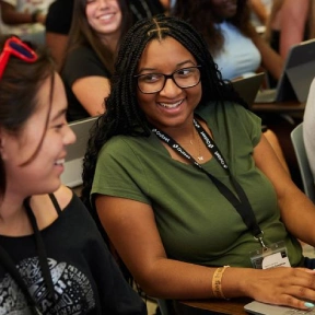 Two students are sitting and conversing in a classroom. One wears glasses and a green shirt, both have lanyards. Laptops and other people are visible in the background.