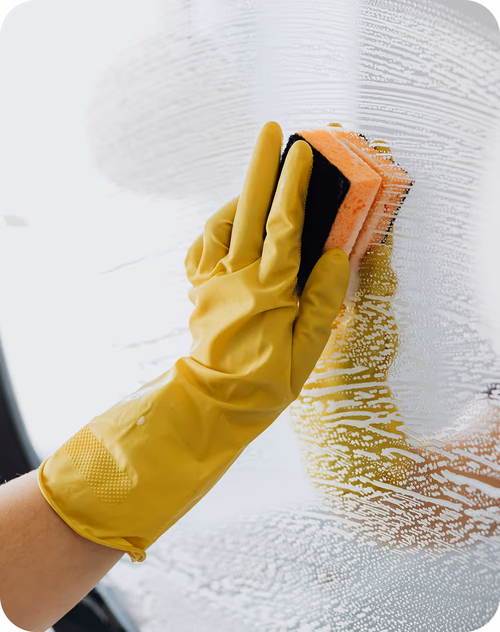 Hand wearing a yellow glove scrubbing a glass surface with a sponge covered in soap suds.