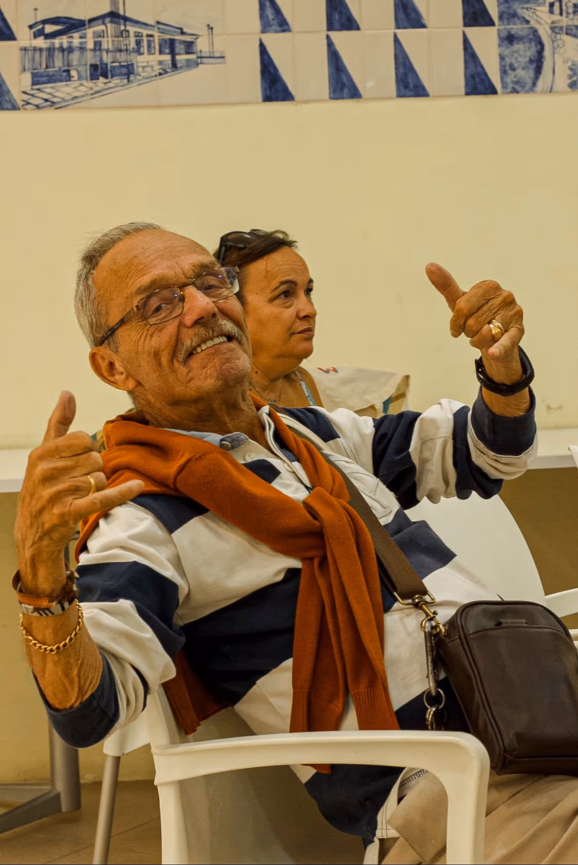 Idoso sorridente com óculos e camisola às riscas sentado numa cadeira branca a fazer o gesto de shaka com as duas mãos.