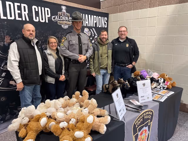 Five people, including a Pennsylvania State Police officer in uniform, stand behind tables filled with teddy bears and informational materials inside a sports venue with a Calder Cup Champions banner.