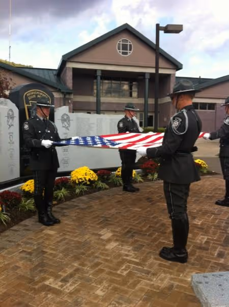 Uniformed officers folding an American flag in a ceremony in front of a memorial wall with flowers.