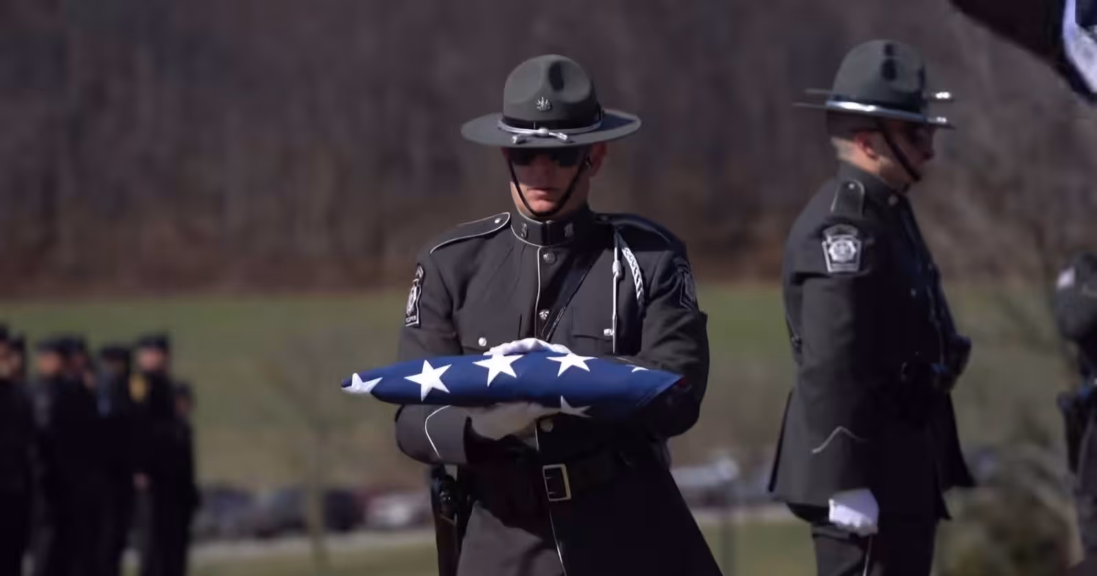 Uniformed officer holding folded American flag during a ceremonial event with other officers present.