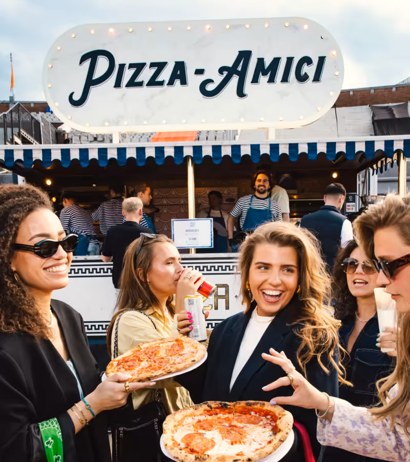Group of five smiling young people enjoying pizza slices in front of a food stall with a sign reading 'Pizza Amici'.