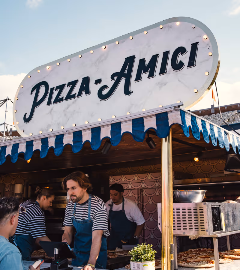 Food stall with sign 'Pizza-Amici' and workers serving customers.