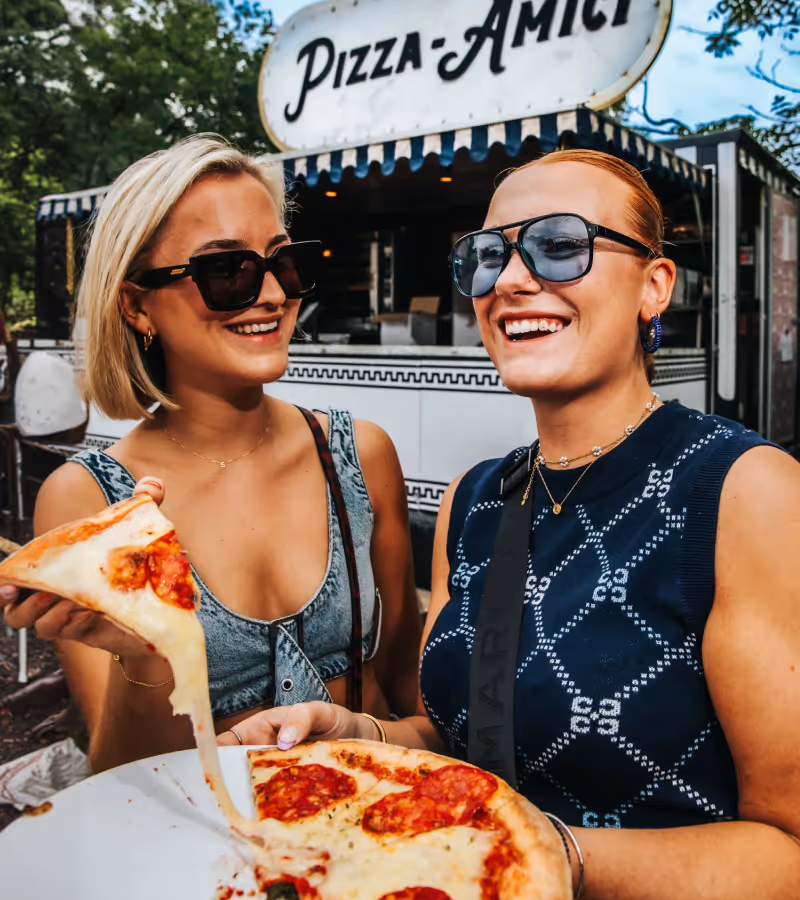 Two smiling women wearing sunglasses holding and sharing a pepperoni pizza slice in front of a food stand with a Pizza-Amici sign.