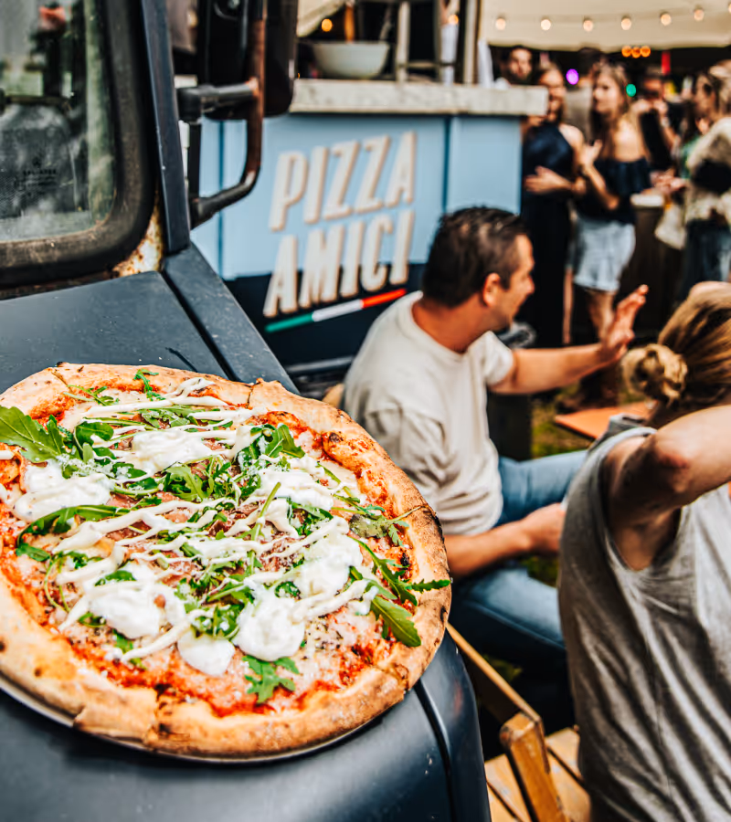 Pizza with arugula and white sauce on a plate resting on a vehicle hood, with people socializing near a food truck labeled Pizza Amici in the background.