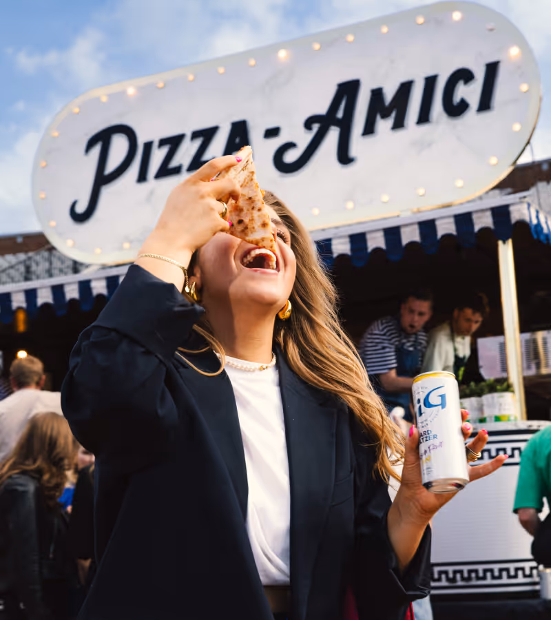 Woman enjoying a slice of pizza and holding a can of hard seltzer in front of a pizza stand named Pizza Amici.