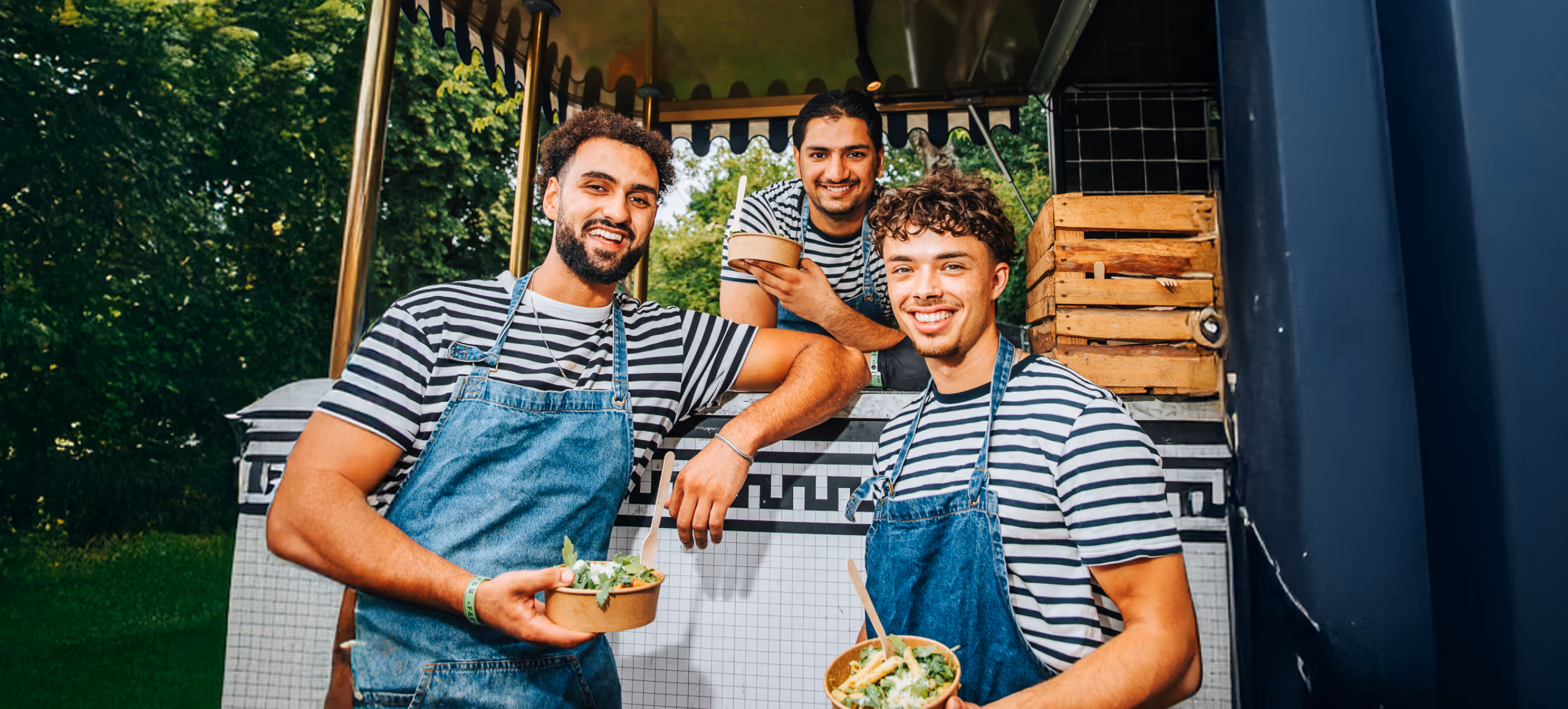 Drie jonge mannen in gestreepte shirts en denim schorten glimlachen terwijl ze buiten gezonde maaltijden in borden vasthouden.