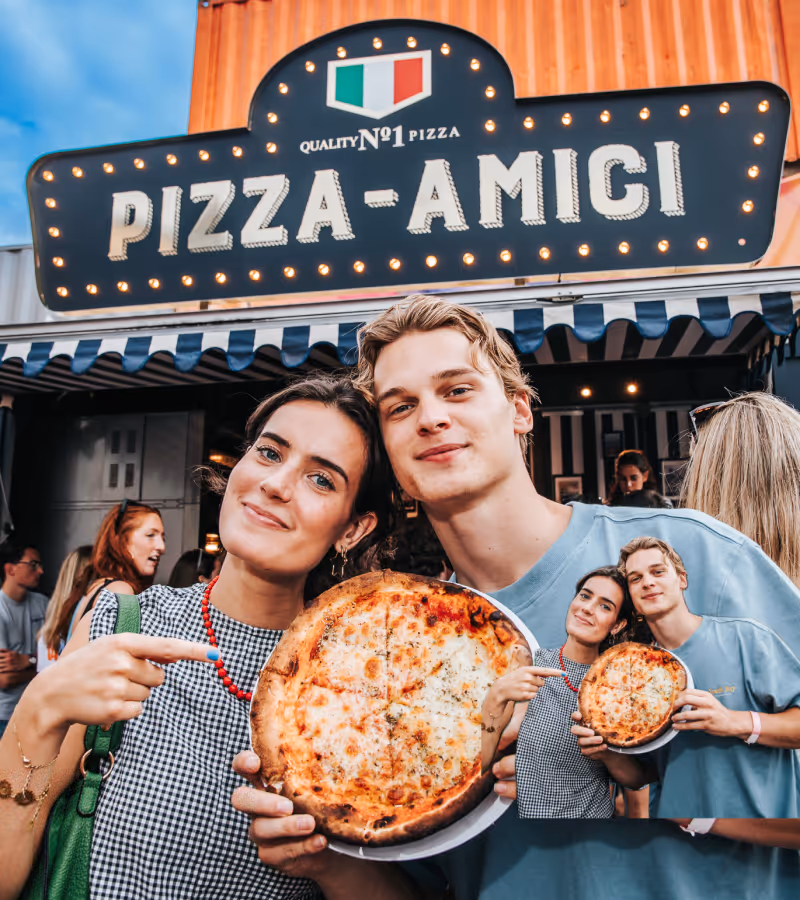 Een glimlachend jong stel toont een pizza buiten pizzeria Pizza-Amici met een bordes en Italiaans vlaglogo.