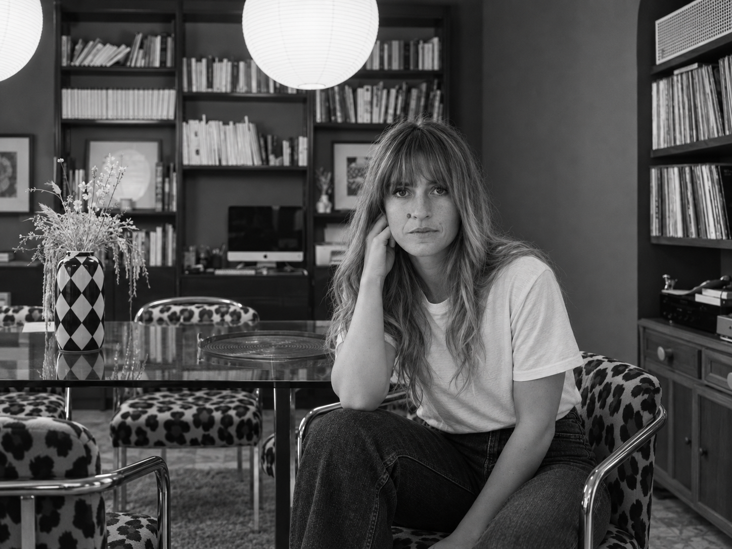 Donna con capelli lunghi e frangia, in maglietta bianca e jeans, seduta su una sedia con rivestimento a motivo maculato in una stanza con librerie, un computer e un tavolo con un vaso decorato e fiori.