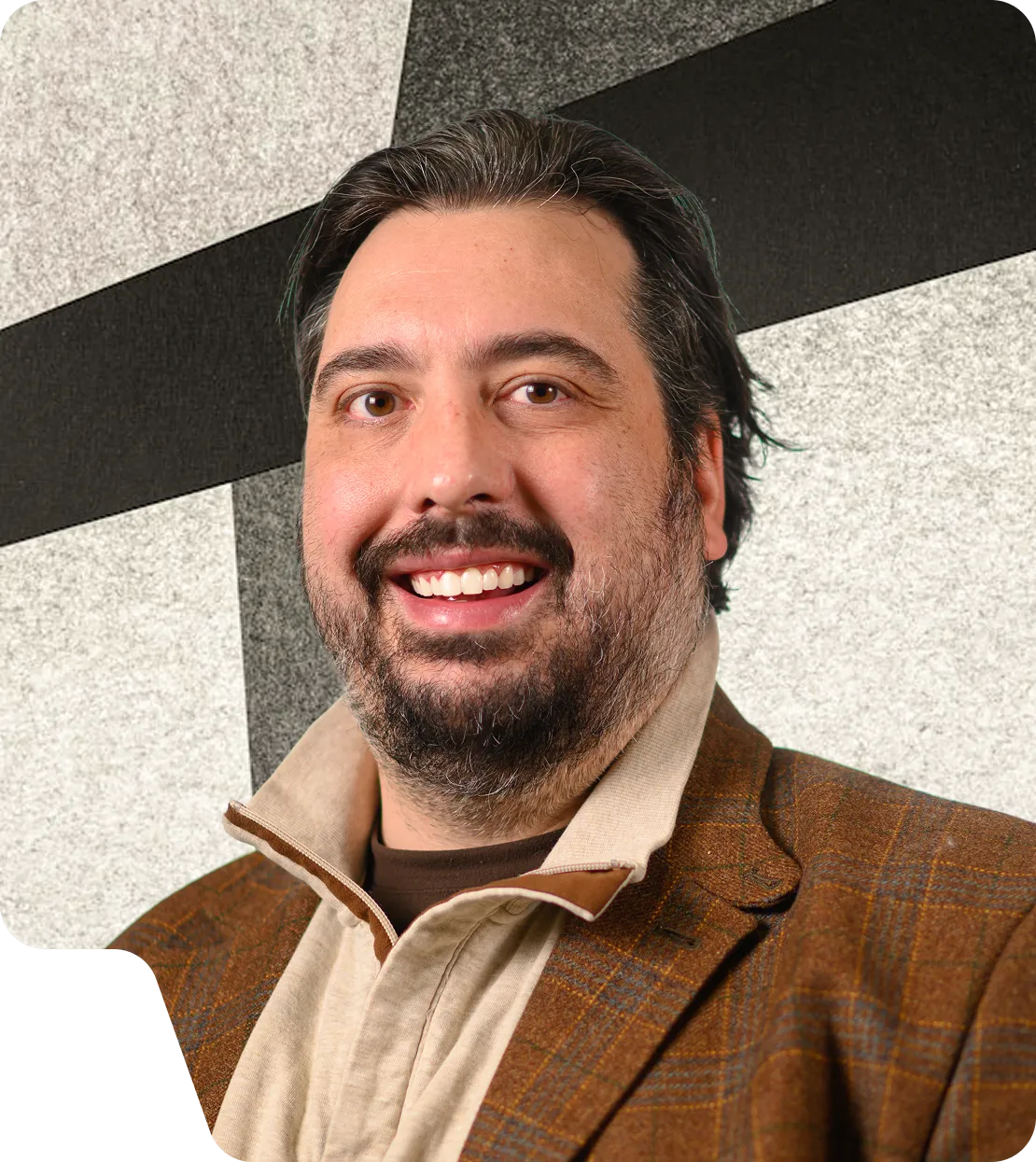 Smiling man with brown hair and beard wearing a beige shirt and brown plaid jacket against a geometric black and white background.