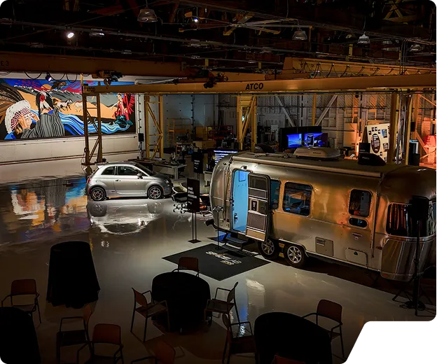 Dimly lit large indoor space featuring a silver Airstream trailer with its door open, a small silver car, chairs, tables, and a colorful mural on the wall.