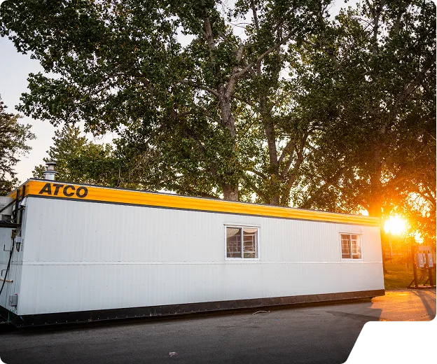 Corner of a gray and yellow ATCO portable building with trees and sunlight in the background.