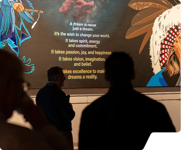 Silhouettes of people in front of a mural featuring an Indigenous figure in a feathered headdress and a motivational quote about dreams and commitment.