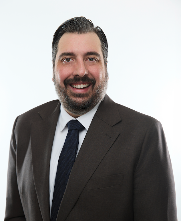 Smiling man with brown hair and beard wearing a beige shirt and brown plaid jacket against a geometric black and white background.