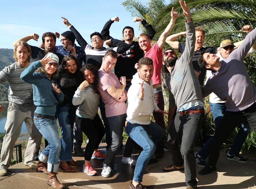 Group of sixteen people outdoors posing energetically with flexed arms and playful gestures against a backdrop of trees and hills.