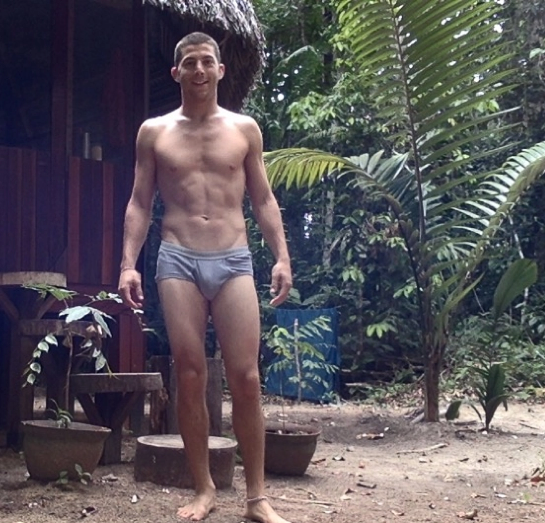 A shirtless Josh wearing gray swim trunks standing outdoors on dirt ground with tropical plants and wooden structures around.