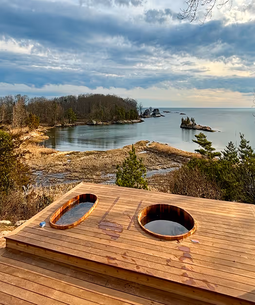 Wooden deck with two round hot tubs overlooking a coastal landscape with trees, rocky shoreline, and a calm sea under a cloudy sky.