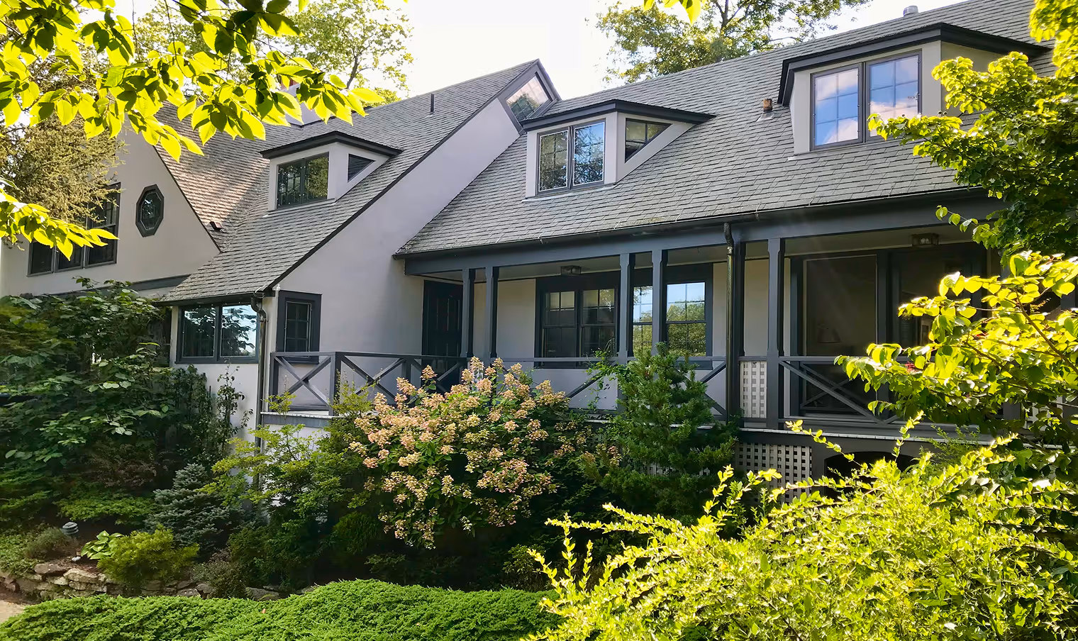 Large house with gray roof, dormer windows, porch, and lush green garden with flowering bushes in front.