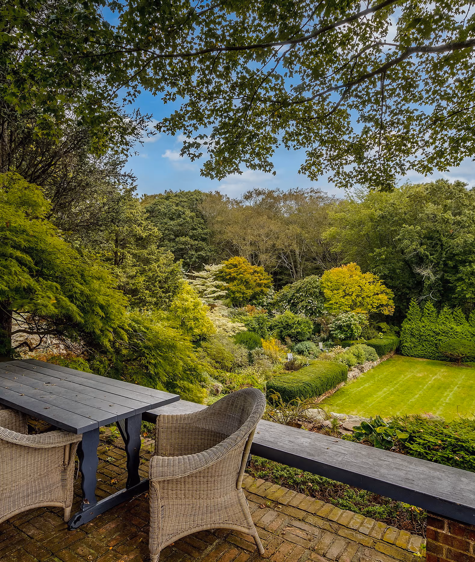 Outdoor wicker chairs and a wooden table on a brick patio overlooking a lush garden with various trees and neatly trimmed hedges.