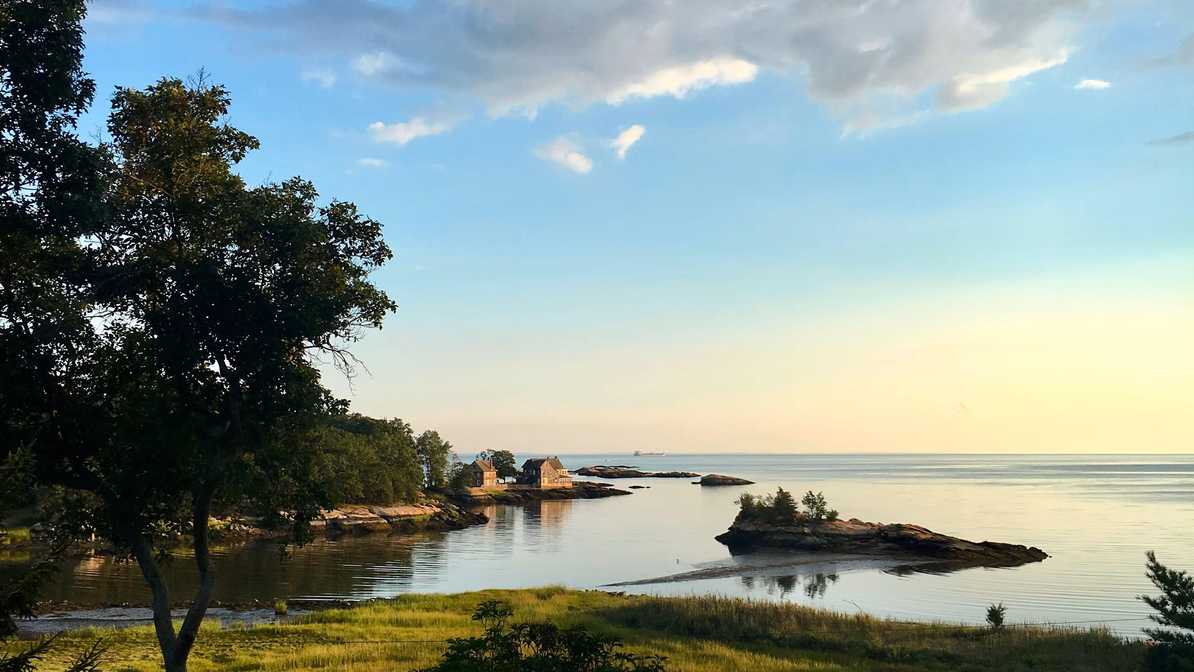 View of a rocky beach with waves crashing and green hills under a partly cloudy blue sky.