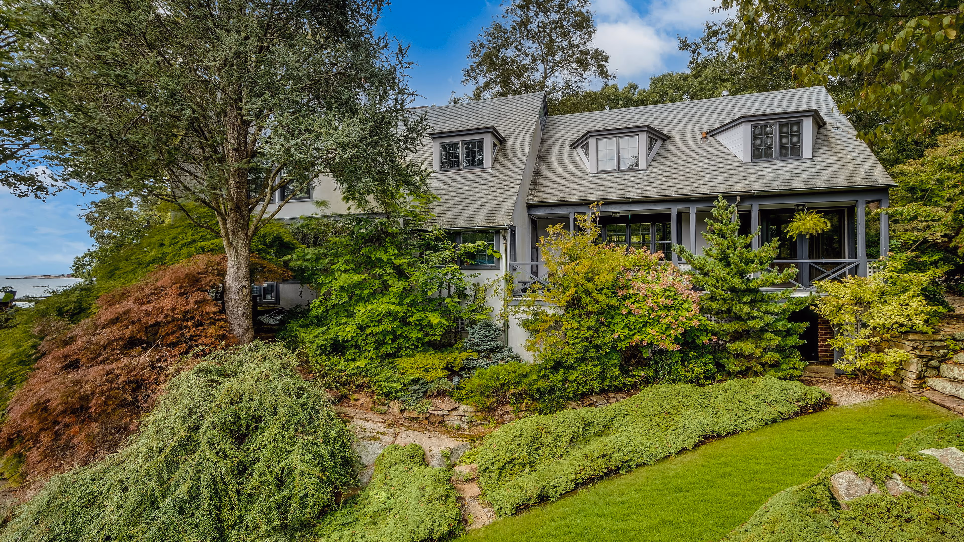 Gray house with a sloped roof surrounded by lush green shrubs, trees, and well-maintained landscaping.