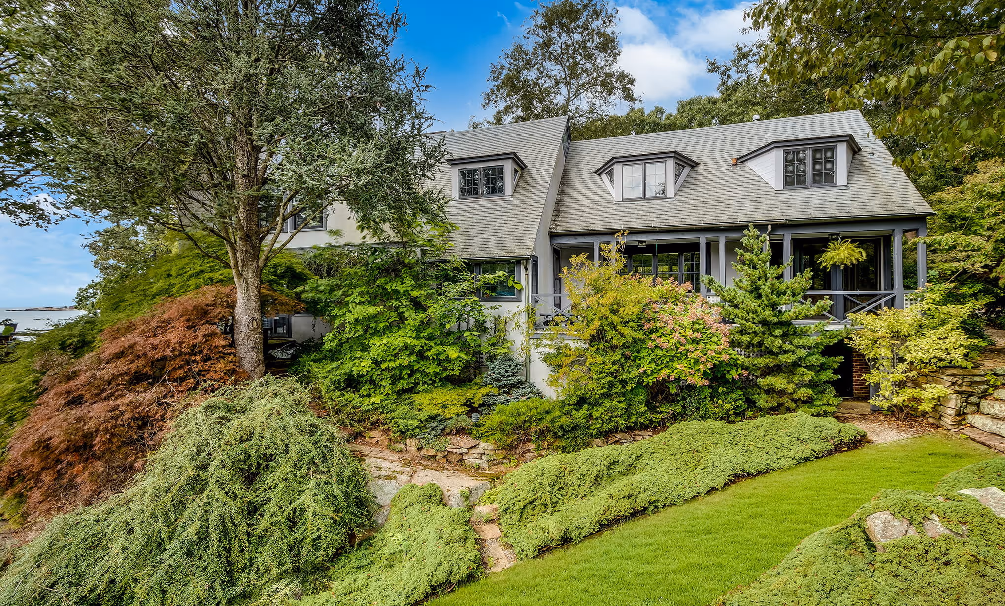Gray house with dormer windows surrounded by dense green and autumn-colored shrubs and trees under a partly cloudy blue sky.