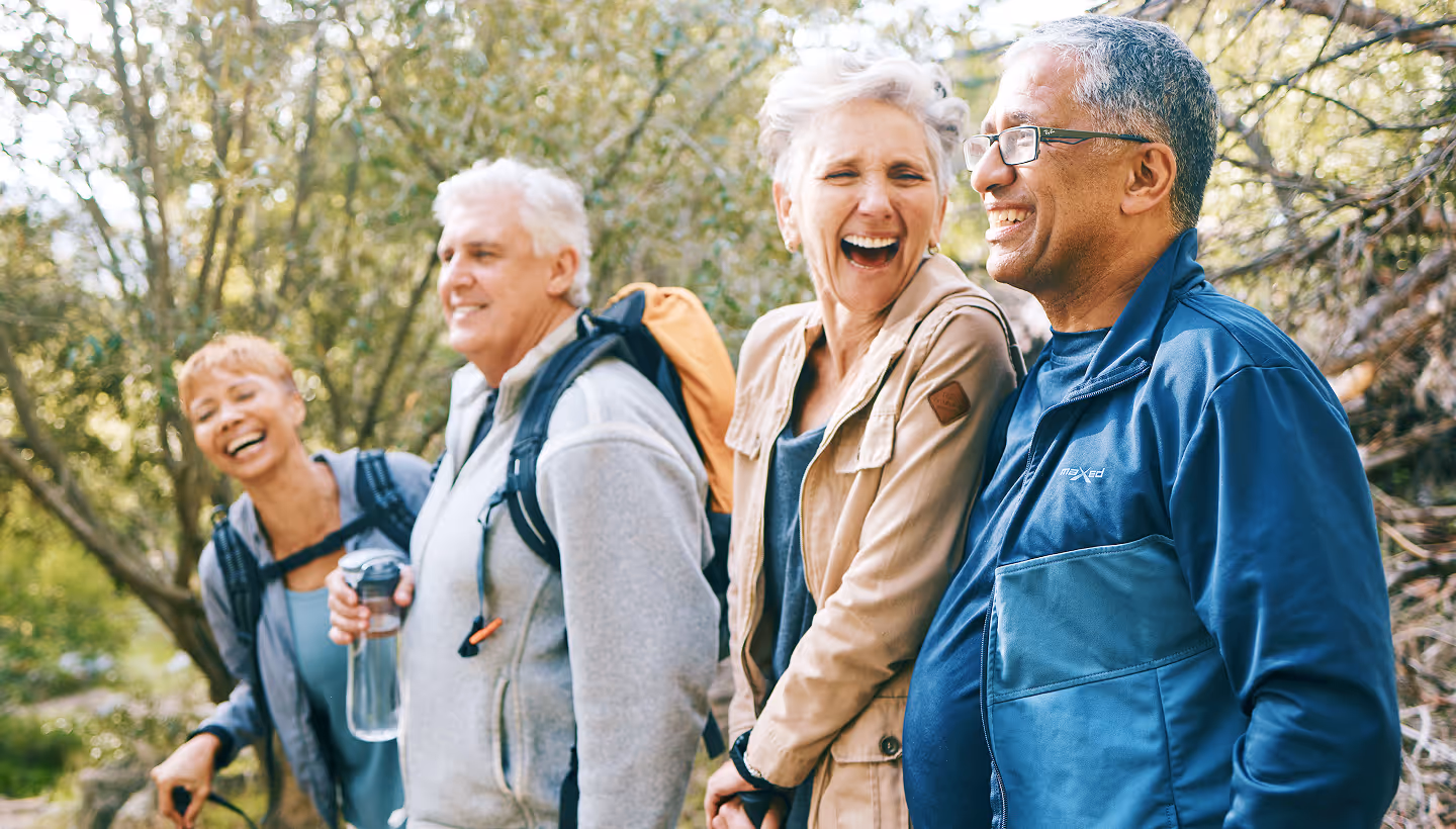 Group of four mature adults wearing outdoor gear and backpacks, smiling and laughing on a forest hike.