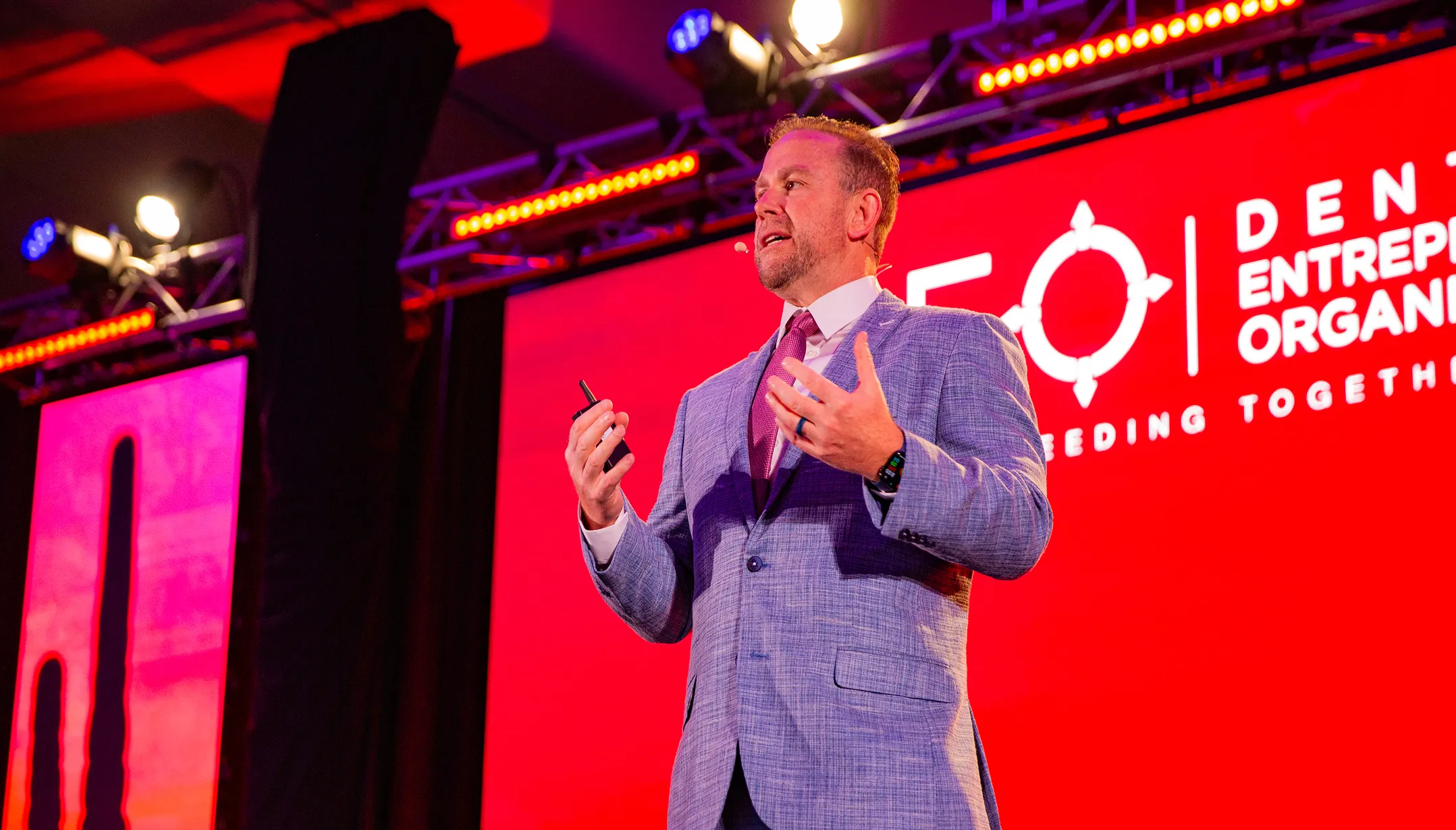 Man in a blue suit speaking on stage with a red background screen