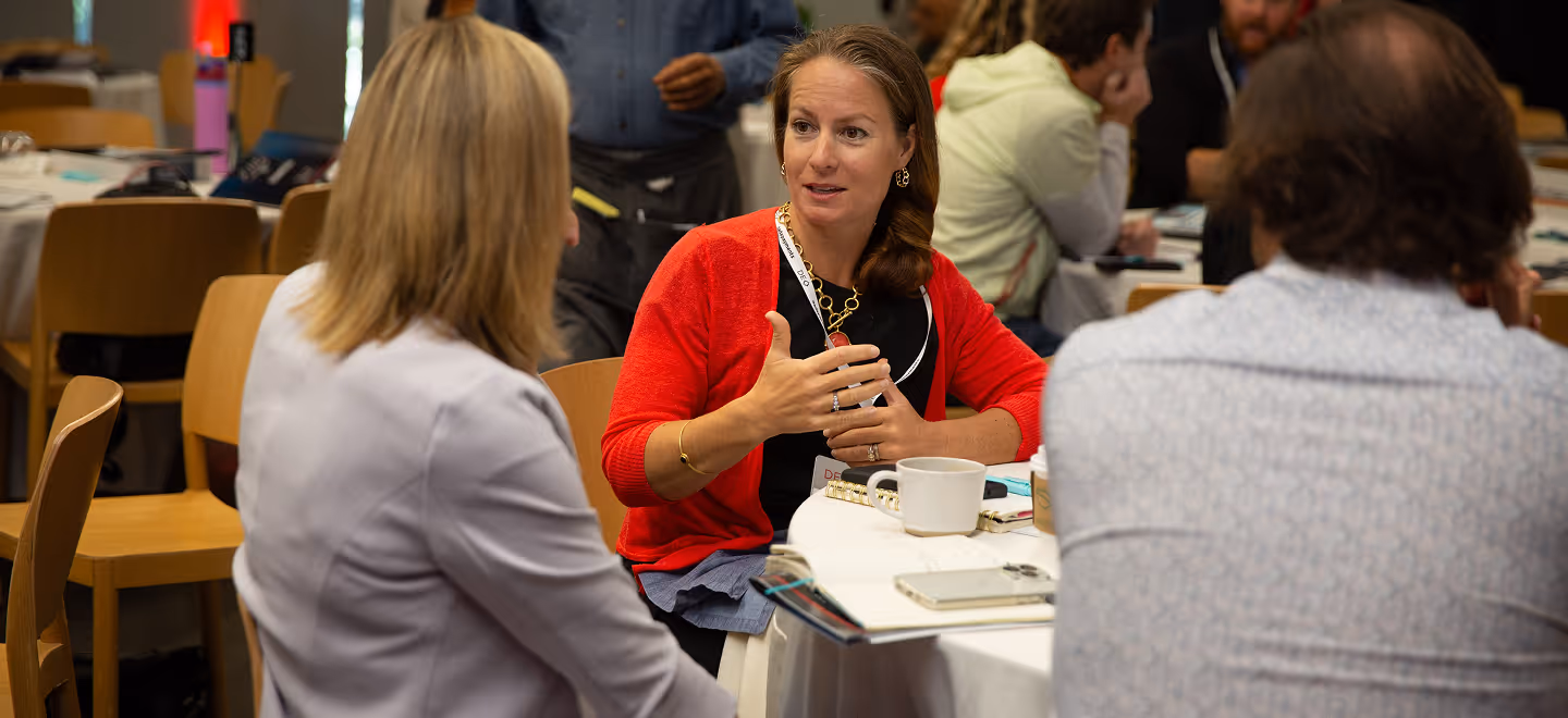 Three people seated at a round table engaged in conversation in a meeting or conference setting.