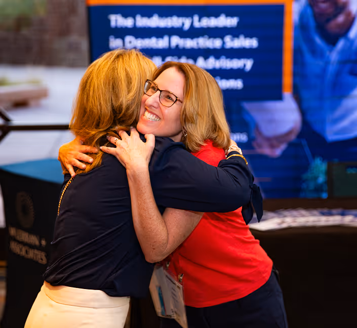 Two women warmly hugging each other indoors at an event, with a display board in the background.
