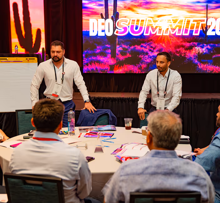 Two men in white shirts standing and speaking to a group seated around a round table during a summit event with a desert sunset background on the screen.