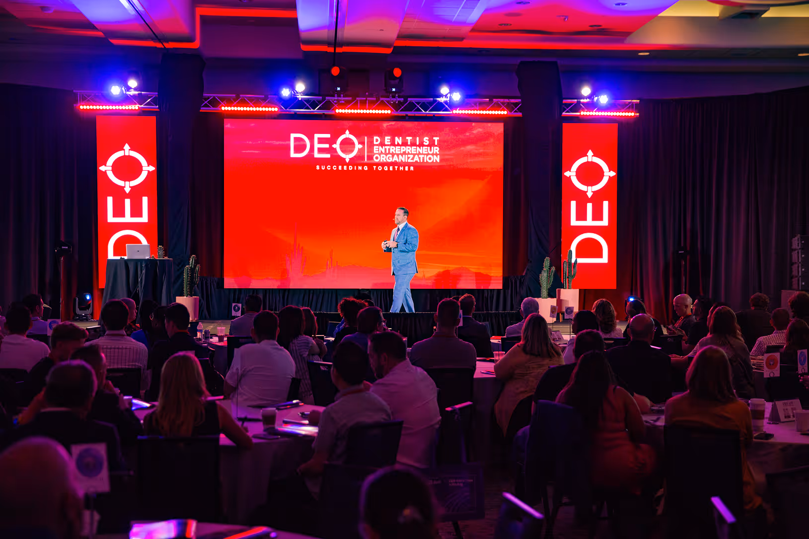 Conference room with a speaker on stage presenting to an audience, backdrop screen displays Dentist Entrepreneur Organization logo and message.